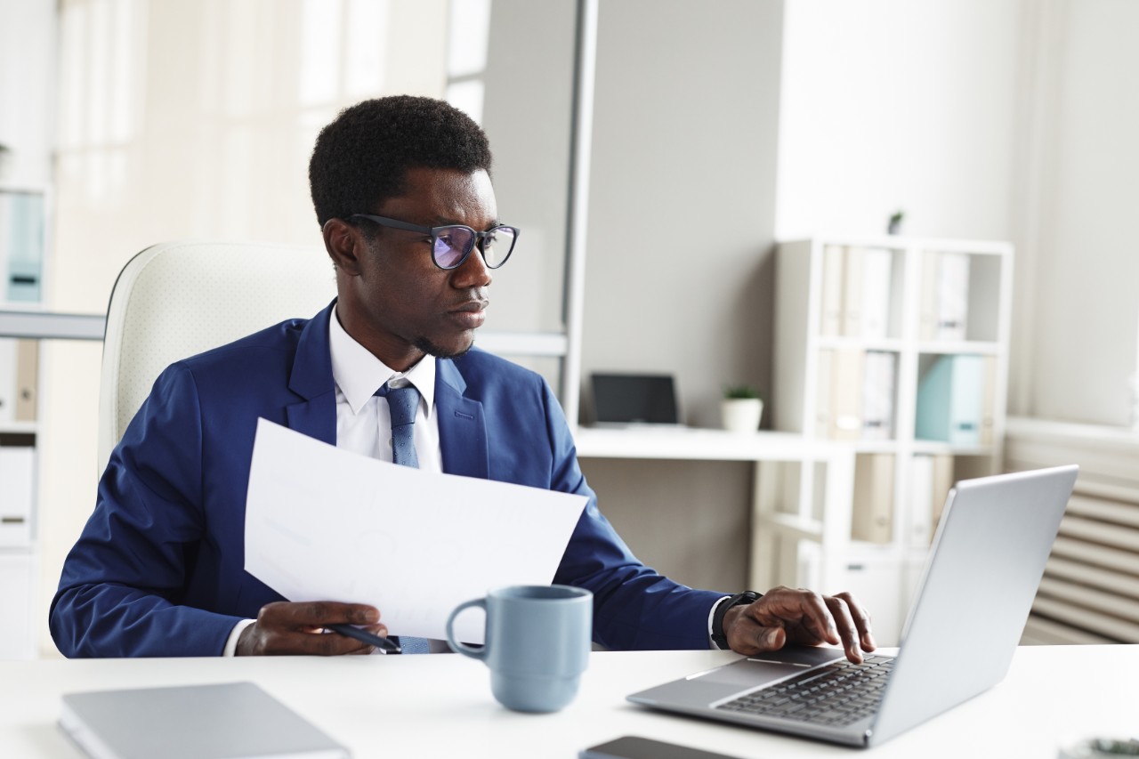 A man sits at a desk holding a document and working on a laptop