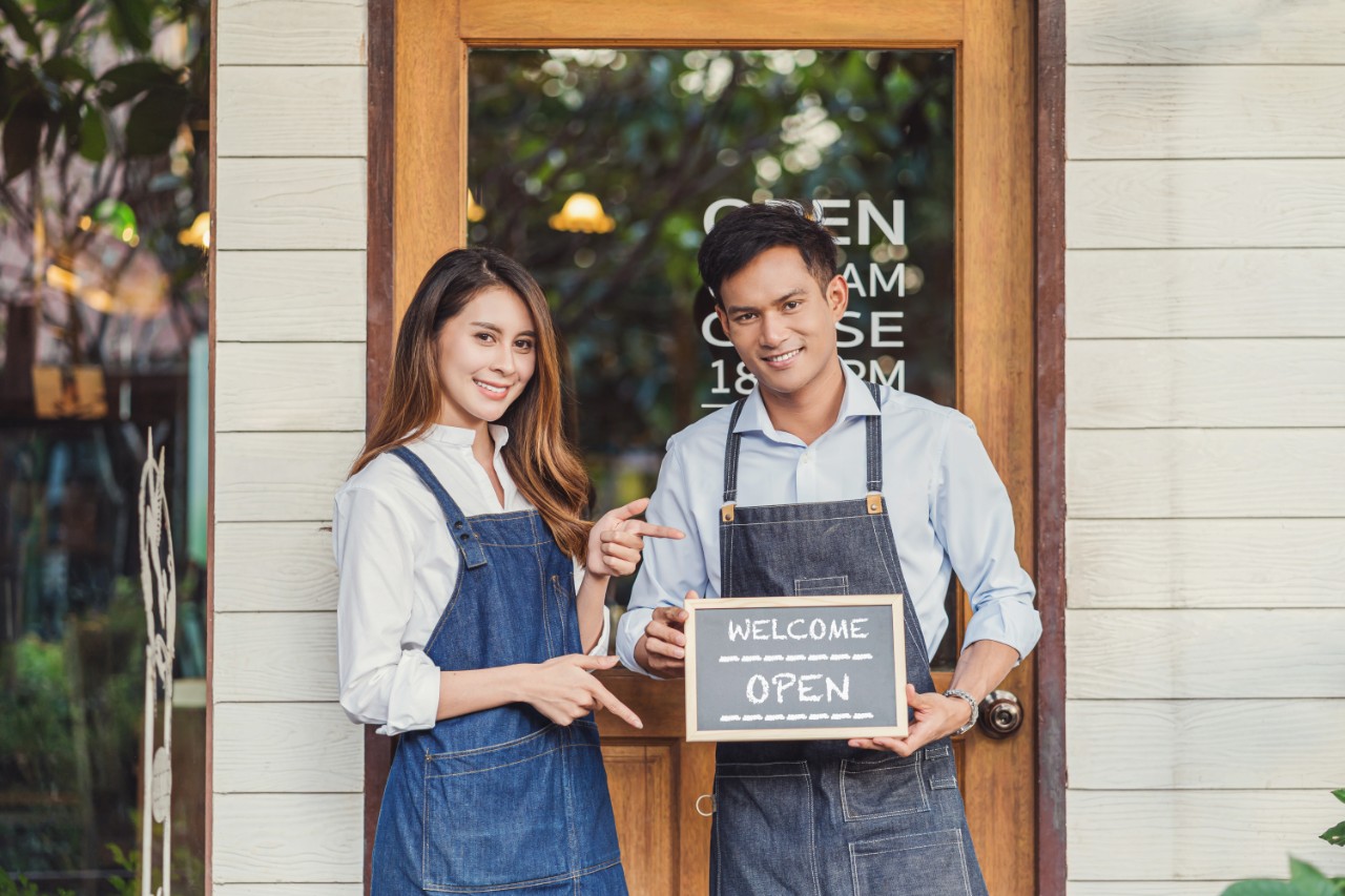 A man and woman stand in front of a storefront holding a sign that says "Open"